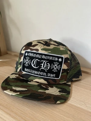Keps Chrome Hearts - Säljer en camouflagemönstrad keps från Chrome Hearts med svart och vit patch framtill där det står 'CHROME HEARTS HOLLYWOOD, USA'. Kepsen har mesh på sidorna och justerbar snapback. Perfekt för dig som gillar streetwear och vill sticka ut.