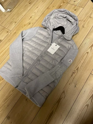 Ljusgrå dunväst med huva från Moncler - Snygg ljusgrå dunväst från Moncler med stickade ärmar och huva. Västen har dragkedja framtill, ribbade muddar och en Moncler-logga på ärmen. Perfekt för lager-på-lager och riktigt trendig look.