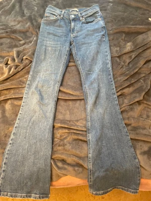 Blå bootcut jeans med hög midja - Säljer ett par klassiska blå bootcut jeans med hög midja och fem fickor. Jeansen har en snygg tvätt och vida ben som ger en retrovibe. Tillverkade i denim med bälteshällor och knappgylf. Perfekta för dig som gillar en avslappnad men trendig stil.