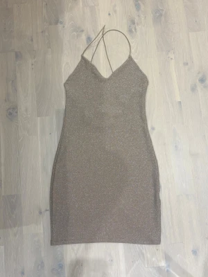 Glittrig beige kort klänning - Säljer en glittrig beige kort klänning med tunna axelband som korsas i ryggen. Klänningen har en figurnära passform och v-ringning framtill. Perfekt för dig som vill sticka ut med en stilren och trendig look.