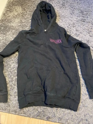 Svart hoodie med rosa Wallride-tryck - Säljer en svart hoodie med huva och känguruficka framtill. På bröstet finns ett rosa Wallride-tryck som ger en cool streetkänsla. Hoodien har ribbade muddar vid ärmslut och nederkant. Perfekt för dig som gillar en avslappnad och stilren look.