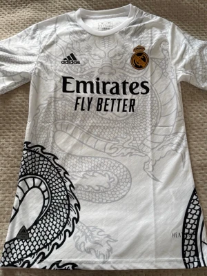 Real Madrid draktröja Adidas vit - Säljer en vit Real Madrid matchtröja från Adidas med ett stort drakmönster i grått över hela tröjan. Tröjan har klubbmärke och Adidas-logga på bröstet samt Emirates Fly Better-tryck. Tillverkad i lätt och funktionellt material, perfekt för fotboll eller träning.