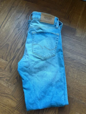 Ljusblå jeans från Jack & Jones - Snygga ljusblå jeans från Jack & Jones med klassisk femficksdesign. Jeansen har en läderpatch med logga bak i midjan och subtila slitningar framtill. Perfekta för en avslappnad och trendig look. Storlek W28 L32