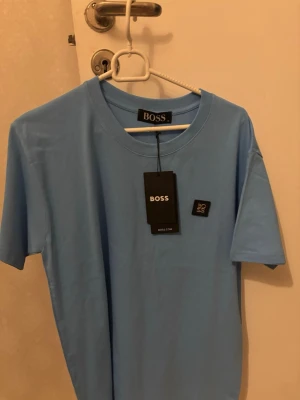 Ljusblå t-shirt från BOSS - Snygg ljusblå t-shirt från BOSS med klassisk rund halsringning och diskret logotyp på bröstet. Tillverkad i mjuk bomull för en bekväm känsla. Perfekt till jeans eller shorts för en clean och stilren look.