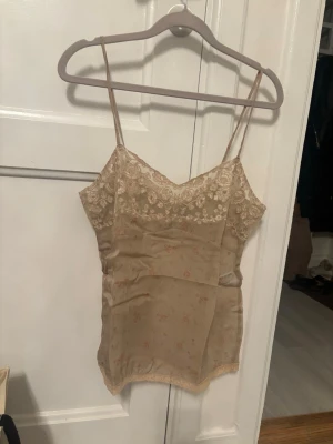 Beige spetsdetaljerad linnetopp - Säljer en beige linnetopp med tunna axelband och v-ringad spetskant upptill. Toppen har ett diskret blommigt mönster och är gjord i ett tunt, lätt material som känns lyxigt och somrigt. Perfekt att styla med jeans eller kjol.