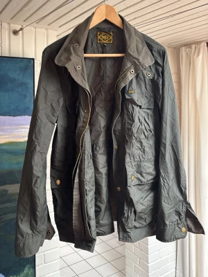 Field jacka   - Snygg mörkgrön vindjacka från Obey med flera fickor framtill, dragkedja och tryckknappar. Jackan har klassisk krage och justerbara ärmslut. Tillverkad i lätt, vindtätt material som passar perfekt för blåsiga dagar.