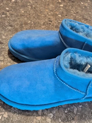 Blåa fluffiga boots i mocka från UGG - Säljer ett par coola blåa boots från UGG med mjukt fluffigt foder och ovandel i mocka. Skorna har en enkel design med rund tå och låg skaft, perfekta för att hålla fötterna varma. Snygga sömmar och matchande blå sula ger en clean look.
