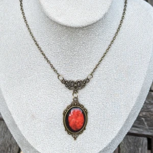 Rött Kristallhänge Halsband - Detta vackra halsband har ett rött kristallhänge som fångar ljuset på ett förtrollande sätt. Den djupa röda stenen ger en elegant och mystisk touch, perfekt för att komplettera din gotiska stil. Material: Ospecificerat Mycket fint skick 