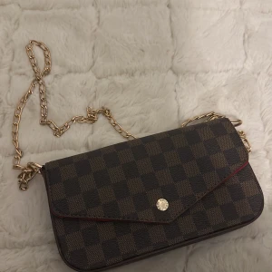 Brun och svart rutig axelväska med kedja - En elegant axelväska med ett rutigt mönster i brunt och svart. Väskan har en guldfärgad kedja som axelrem och en knappstängning. Insidan är röd och har flera fack för förvaring.