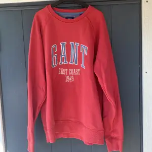 En röd sweatshirt från GANT med texten 'GANT EAST COAST 1949' tryckt på framsidan. Tröjan har långa ärmar och ribbstickade muddar vid ärmslut och nederkant.