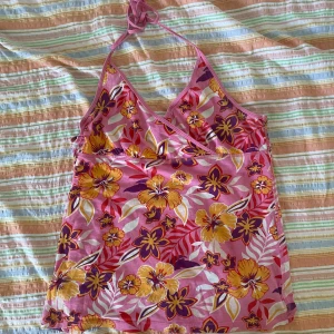 Blommig halterneck topp - En färgglad blommig halterneck topp med rosa, gula och lila blommor. Toppen har en omlott-design framtill och smockdetaljer på baksidan. Den är ärmlös och knyts runt nacken.