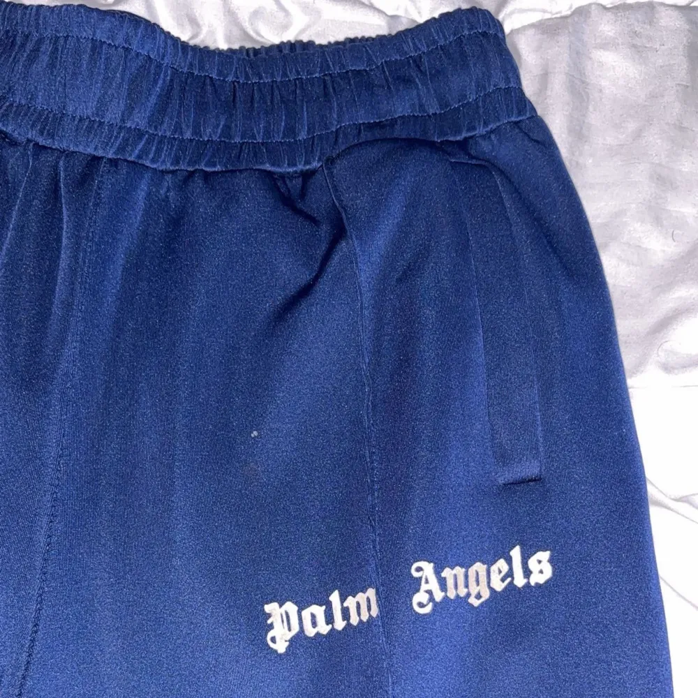 Ett par mörkblå mjukisbyxor från Palm Angels med vit text på vänster ben. Byxorna har en elastisk midja och vita ränder längs sidorna. De har även dragkedjor vid bensluten.. Jeans & Byxor.