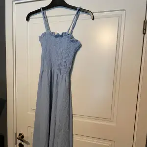 En ärmlös ljusblå klänning i trikå. Klänningen är knälång och har justerbara axelband samt ett smockat liv med volang längst upp.   -nypris 149 SEK