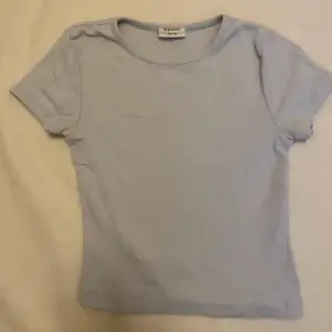 En ljusblå t-shirt med små ränder i tyget. Från KappAhl och storlek 146-152. Ganska kort i magen, men inte riktigt lika kort som en magtröja.