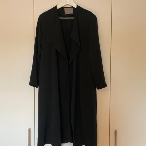 Svart tunn kappa - Svart tunn kappa från Vero Moda i storlek XS, band medföljer som kan sättas vid midjan om man så önskar. Den räcker ner till början av vaderna ungefär på mig som är 165 cm lång 