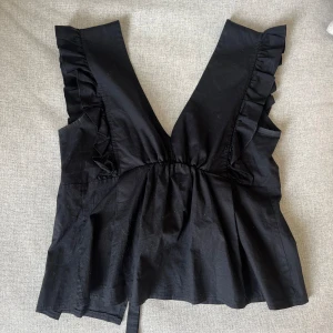 Topp  - Svart topp. Med fina detaljer vid axeln och knyte i ryggen. Knyter i ryggen går att justera. Nästintill oanvänd. 