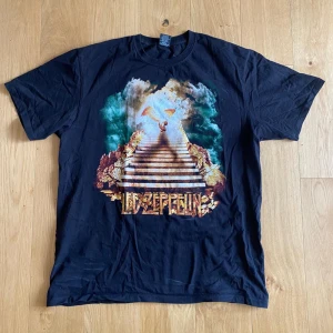 Svart Led Zeppelin t-shirt - En svart t-shirt med Led Zeppelin-tryck på framsidan och baksidan. Framsidan har en färgglad illustration med en trappa och en ängel, medan baksidan har bandets namn och en cirkulär logotyp. T-shirten är av märket Rock Yeah och är i storlek XL.