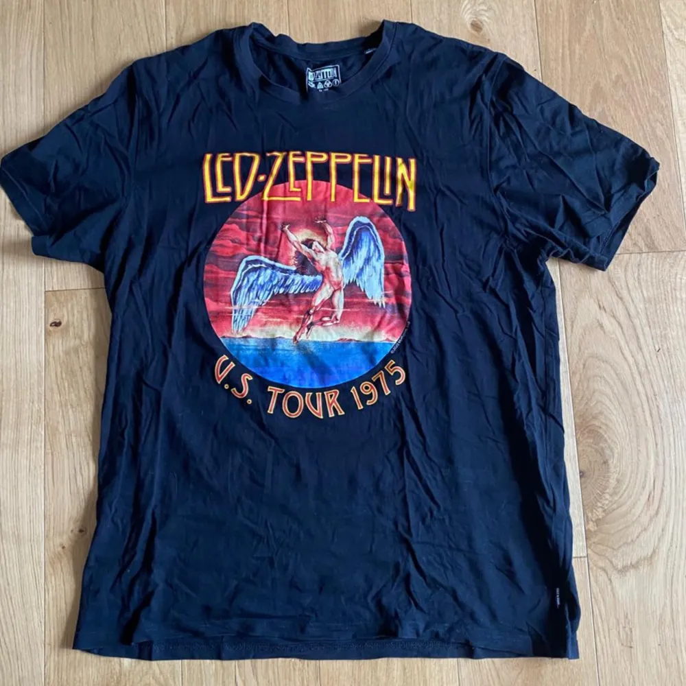 En svart T-shirt med Led Zeppelin-tryck på framsidan. Trycket visar en illustration och texten 'U.S. Tour 1975'. T-shirten är kortärmad och tillverkad av bomull.. T-shirts.