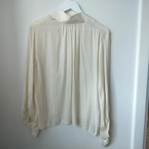 Cremevit underbar blus med hög krage och knyt - Superfin classy blus med hög krage och knyt baktill, ärmarna är voluminösa och flowy. Använd vid ett tillfälle! Från Wera/Åhlens, köpt för 400 eller 500 kr.
