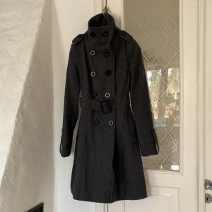 Ullkappa från Vila, mörkgrå med skärp, strl xs - Ullkappa från Vila i fin mjuk kvalitet, 60% ull och 40 % polyester. Dubbelknäppt med skärp. Fina detaljer i ärmslut.  Längd: 92cm Mycket fint skick