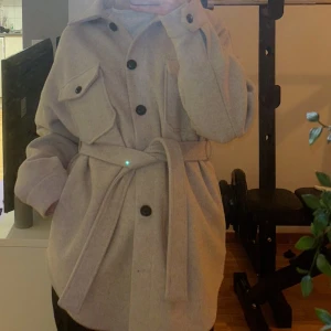 Beige kappa (schacket) - En beige kappa från nelly! Slutsåld, går inte att köpa längre (originalpris 699kr). Storlek 32 men stor i storleken, sitter mer som en 34/36. Använd några gånger, men är i väldigt fint skick! Säljer då den inte kommer till användning längre