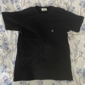 Acne studios smiley face-t-shirt - Svart smiley t-shirt från acne studios men köpt second hand, lite mer slim i modellen (andra bilden). Superfin men jag gillar mer oversize t-shirts✨ Pyttelitet hål på ena axeln (tredje bilden) men det syns knappt när man har på sig den :) 