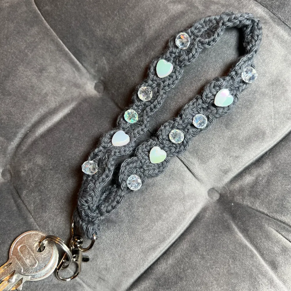 Egenvirkat nyckelband i mörkgrått bomullsgarn med holografiska hjärtan,  diamantformade pärlor och en silverfärgad  karbinhake.. Accessoarer.