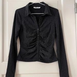 Glittertopp - Glittrig svart tröja från NA-KD i storlek S. Rynkad fram vid knapparna, kan knäppas hela vägen upp till kragen. Stretchigt material. Använd en gång, i fint skick. Bara att fråga om det är något du undrar! Finns fler bilder.