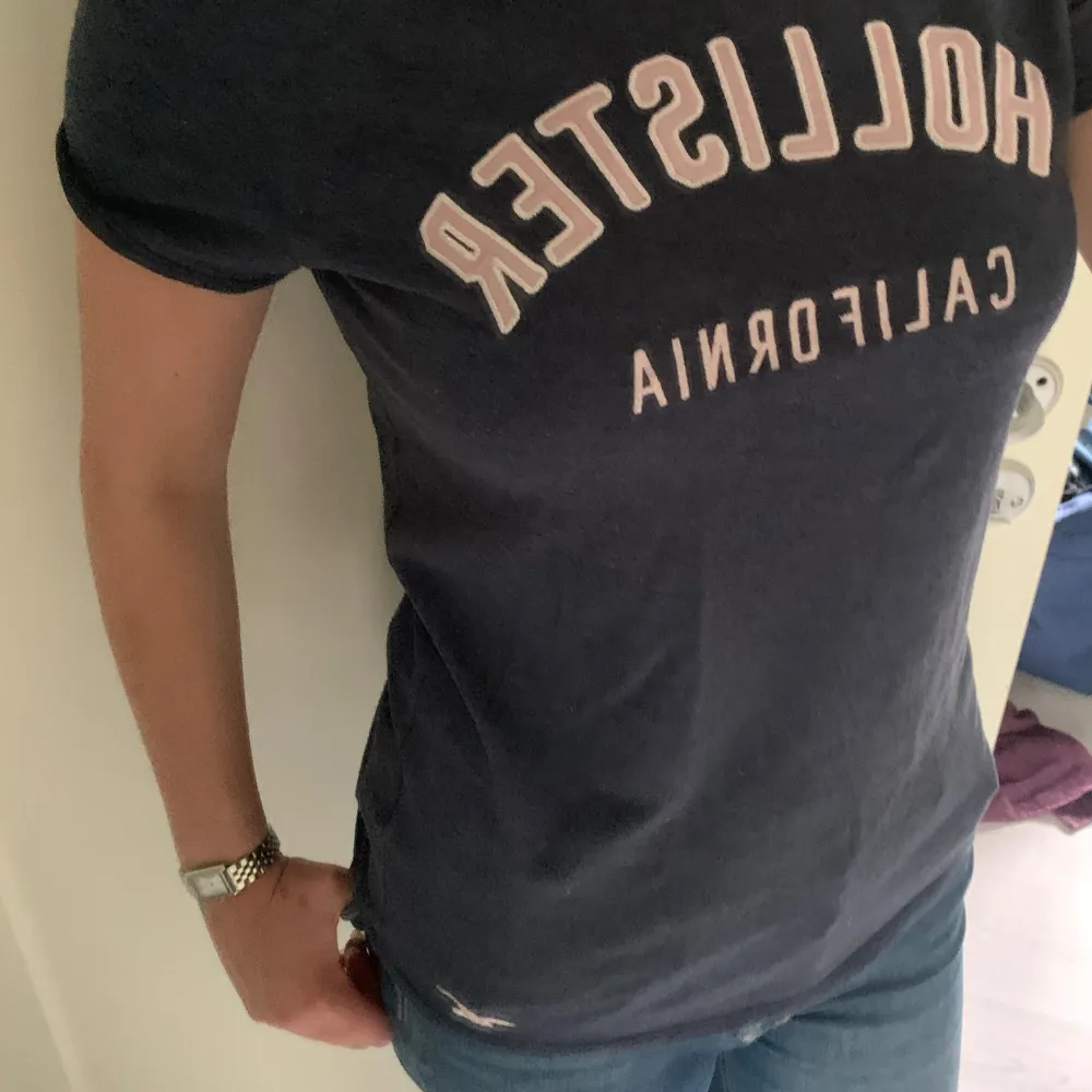 En super gullig Hollister T-shirt med texten ”Hollister California”. Den är Marinblå med rosa/vit text❤️Tröjan passar mig som vanligtvis har S. Bra skick. Skriv vid funderingar!❤️. T-shirts.