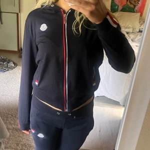 Mörkblått set  - En supersnygg tracksuit/mjukisset. En mörkblå munkjacka med dragkedja framtill och röd-vit randig detalj längs dragkedjan. Hoodien har långa ärmar och Monclers logotyp på bröstet. Mörkblå mjukis byxor med lågmidja och som är 