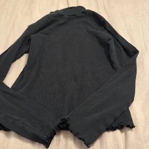 Svart långärmad tröja - En svart långärmad tröja med ribbad struktur och vågiga kanter vid ärmslut och nederkant. Tröjan är i ett enkelt och stilrent utförande.