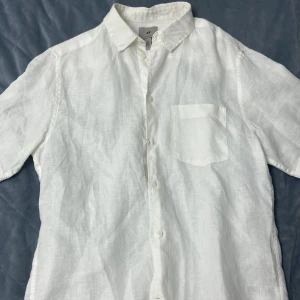 Vit linneskjorta från H&M - En vit linneskjorta från H&M med korta ärmar och en bröstficka. Skjortan har en klassisk krage och knappar framtill. Den är i regular fit och tillverkad av linne.