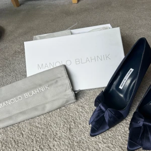Mörkblå pumps från Manolo Blahnik - Äkta eleganta mörkblå pumps från Manolo Blahnik med spetsig tå och rosett framtill. Skorna är i satin och 9 centimerters klack. Använda en gång,  lite smutsiga på sulan. Dustbag och skolåga tillhör. Köparen står för frakt. NYPRIS 9000