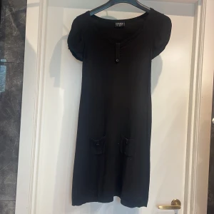 Svart klänning från Soaked in Luxury - En elegant svart klänning från Soaked in Luxury med korta ärmar och en knappdetalj vid halsen. Klänningen har två fickor framtill med knappar. stretch material passar smal. 