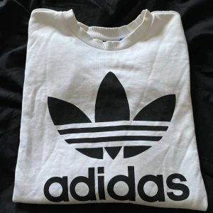 Vit långärmad tröja från Adidas - Säljer en vit långärmad tröja från Adidas med deras klassiska logga i svart på framsidan. Tröjan är i bra skick och perfekt för en avslappnad stil. den har fickor.Den är gjord av bomull och är superbekväm att ha på sig. Passar perfekt till både vardag och träning!