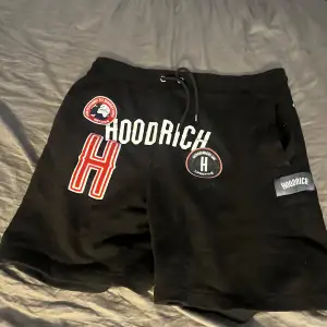 Säljer ett par svarta shorts från Hoodrich. De har coola tryck och broderade detaljer på framsidan, inklusive en stor röd och vit 'H' och flera märken. Shortsen har en dragsko i midjan för justerbar passform och är gjorda av ett bekvämt material. Perfekta för en avslappnad stil!