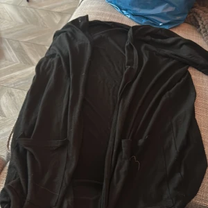 Svart långärmad kappa - Säljer en svart långärmad tröja som är lång. Den är i bra skick. Den har en enkel och stilren design med två fickor framtill. Passar bra till både vardag och fest.