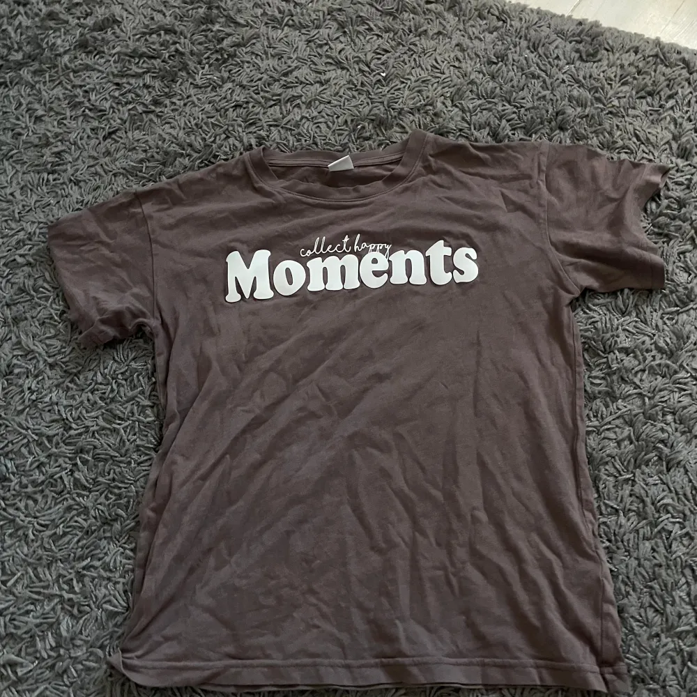 En brun t-shirt från Lindex med texten 'collect happy Moments' tryckt i vitt på framsidan. T-shirten har korta ärmar och är tillverkad i bomull.. T-shirts.