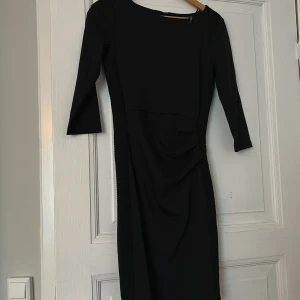 Svart klänning med trekvartsärm - Säljer en elegant svart klänning med trekvartsärm. Klänningen har en stilren design med en liten rynkdetalj på sidan som ger en fin passform. Perfekt för festliga tillfällen eller en kväll ute. Klänningen är i nyskick och har en normal passning.
