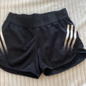 Svarta shorts från Adidas - Ett par svarta shorts/hot pants från Adidas med vita ränder på sidorna. Shortsen har en bred elastisk midja och är tillverkade av ett lätt och andningsbart material. säljer pga att dom är lite försmå 🌟