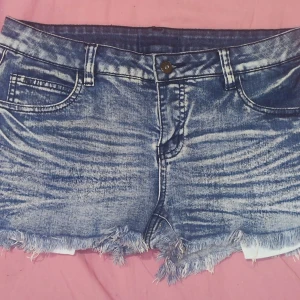 Blå jeansshorts med fransar - Säljer ett par snygga blå jeansshorts med fransar i nederkant. De har en cool tvättad look och är perfekta för sommaren. Shortsen har en knapp och dragkedja framtill samt fickor både fram och bak. Perfekta för en avslappnad och trendig stil!