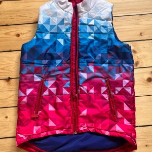 Flerfärgad quiltad väst med dragkedja - Säljer en cool flerfärgad quiltad väst med dragkedja. Västen har ett geometriskt mönster i blått, rött och vitt och är tillverkad av 100% polyester. Den har två fickor med dragkedjor på framsidan och är perfekt för höst och vår. Västen är i bra skick och redo för nya äventyr!