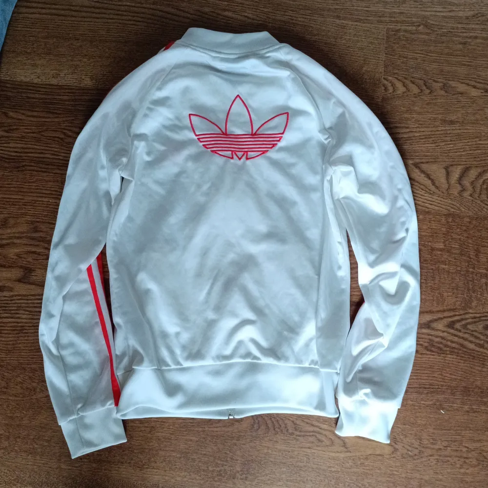 Säljer en snygg vit Adidas zip-up med röda ränder längs ärmarna och Adidas logga på bröstet och ryggen. Jackan har dragkedja framtill och två fickor med dragkedja. Perfekt för en sportig look eller vardagsstil. Passar bra till både träning och fritid.. Jackor.