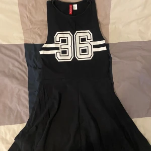 Svart klänning från Divided - Säljer en svart klänning från h&m med ett stort vitt nummer 36 tryckt på framsidan. Klänningen är ärmlös och har en sportig stil med två vita ränder på vardera sida om numret. Perfekt för en avslappnad sommarlook eller en sportig fest! 🖤