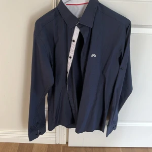 Mörkblå skjorta från Moods of Norway - Säljer en snygg mörkblå skjorta från Moods of Norway. Skjortan är långärmad och har ett diskret randigt mönster. Den är tillverkad av bomull och har knappar längs framsidan. Perfekt för både vardag och fest!