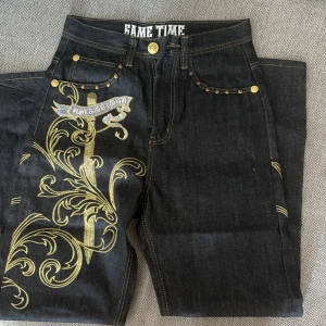 Svarta jeans med guld broderi från Game Time - Säljer ett par svarta jeans från Game Time med snyggt guld broderi. Jeansen har en cool design med detaljer som en broderad svärd och vingar på bakfickorna. De har också guldknappar och nitar💕vet inte om det är äkta