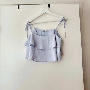 Ljusblå topp från Steph - Säljer en superfin ljusblå topp från Steph. Den är ärmlös med smala axelband som knyts på axlarna och har en volangdetalj framtill. Perfekt för sommaren och kan stylas både till vardags och fest. Toppen är i ett lätt och luftigt material, vilket gör den bekväm att bära under varma dagar.