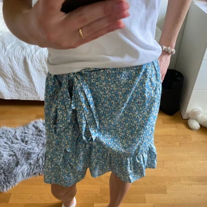 Blommig Kjol - En blå kjol med vita blommor i fint skick! Storlek S och går att knyta i midjan.