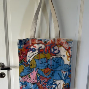 Tygpåse - En cool totebag med smurfarna på! Köpt på  ’Ivan Alexis’ i Köpenhamn för 2 år sedan ☺️ Bra skick men en liten fläck på fickan inuti väskan. Har en liten knapp så man kan knäppa den också. 💚