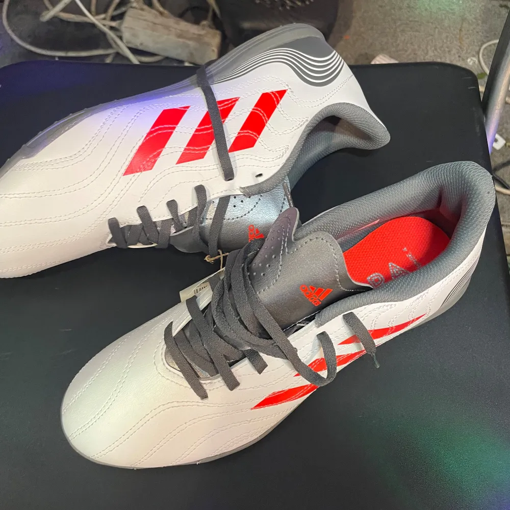 Adidas Copa Sense.4 IN fotbollsskor i grått och rött. Skorna har en slät yta med snörning och är designade för inomhusfotboll. De har en robust sula för bra grepp på inomhusplaner.. Skor.